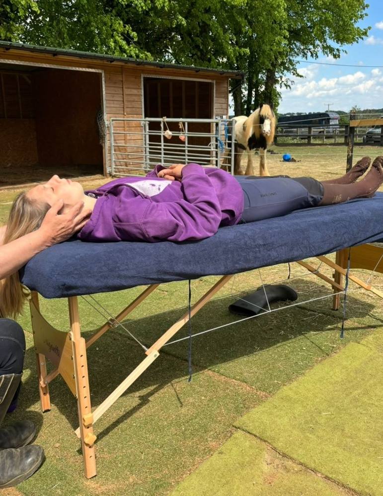 A client relaxing on a treatment table outdoors, with a pony in the background.
